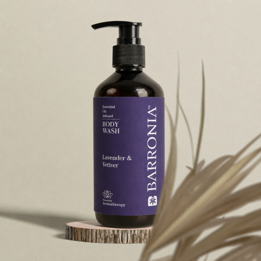 A dark amber bottle of Barronia Lavender & Vetiver infused body wash with a purple label and black pump, displayed on a small wood slice pedestal with dried palm leaves against a neutral beige background.