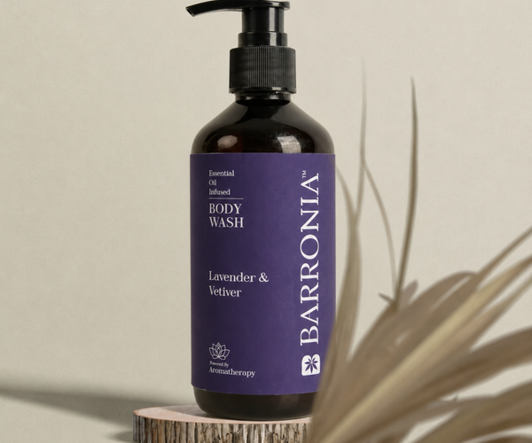 A dark amber bottle of Barronia Lavender & Vetiver infused body wash with a purple label and black pump, displayed on a small wood slice pedestal with dried palm leaves against a neutral beige background.